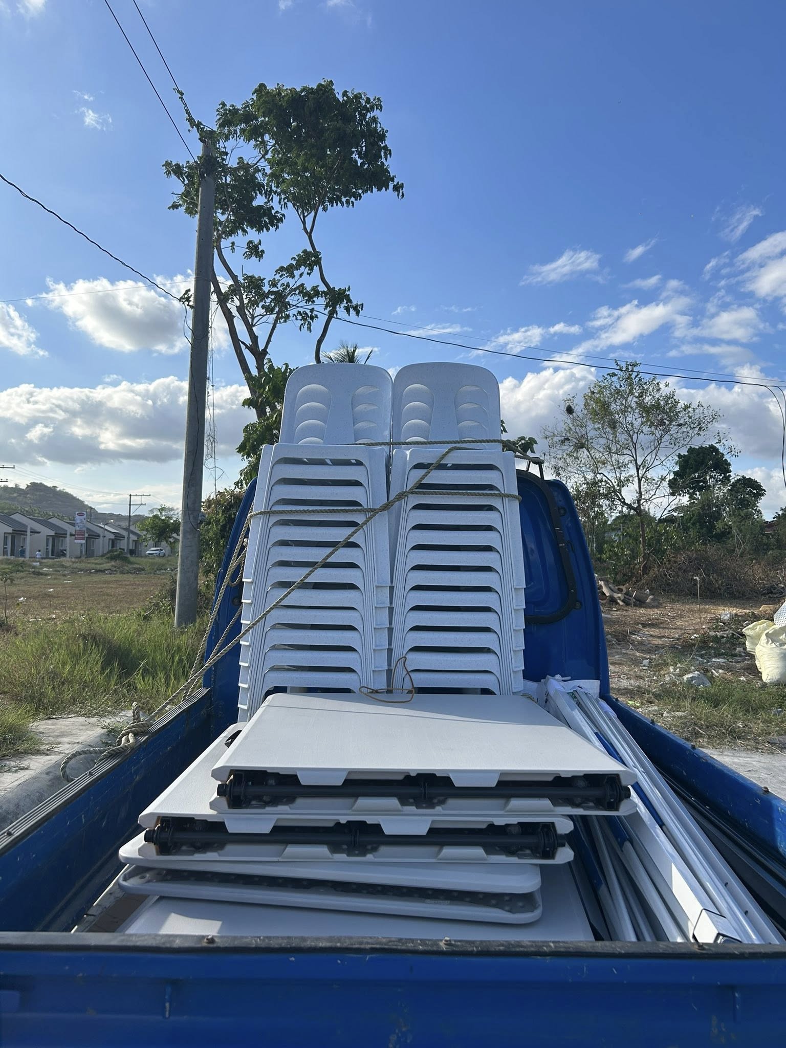 Easy Rental delivery — blue pickup truck bed loaded with stacked white monobloc chairs and folded white tables tied with rope, plus tent frame poles
