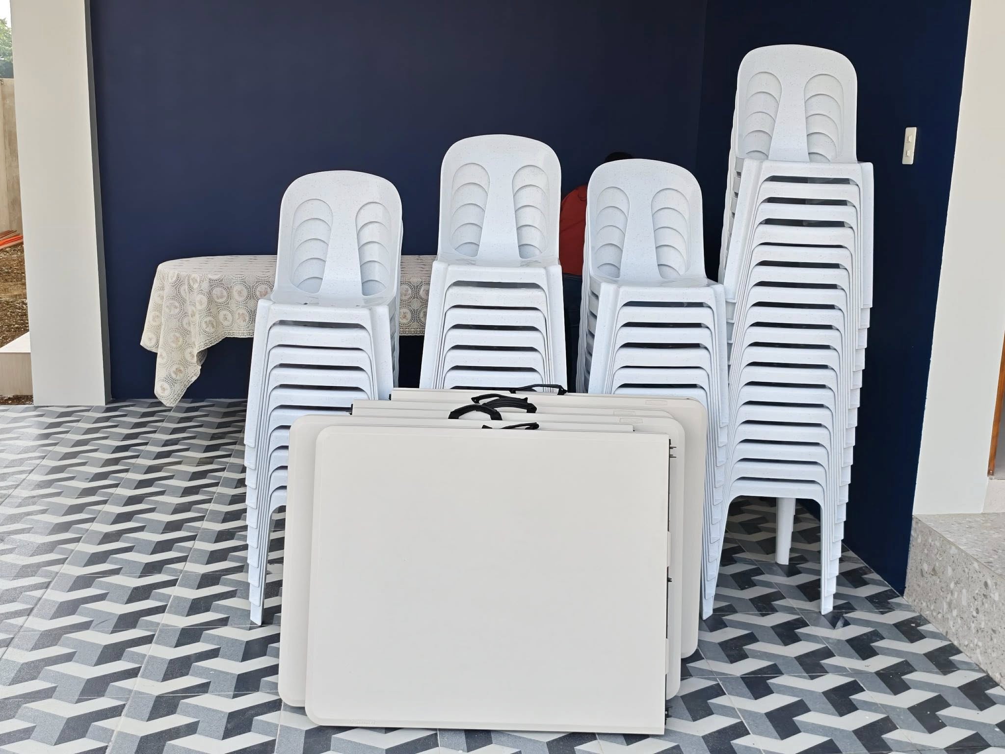 Easy Rental inventory — four stacks of white monobloc chairs and folded white plastic tables staged against a navy wall before dispatch